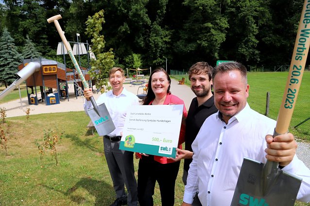 Bild: B&uuml;rgermeister Christian Zwingmann, Klimaschutzmanager Stephan Gunkel, SWL-Gesch&auml;ftsf&uuml;hrerin Evelyn Rudolph und ihr Kollege R&uuml;diger Steinberg (von rechts) bei der symbolischen &Uuml;bergabe des Spendenschecks auf dem Hundeshagener Spielplatz. Die B&auml;ume und Str&auml;ucher im Hintergrund hatte der st&auml;dtische Bauhof bereits im Vorfeld gepflanzt. 